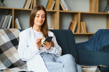 Young happy caucasian woman she 20s in casual clothes hold in hand use mobile cell phone sit on blue sofa indoor rest at home in own room apartment. People lifestyle concept.