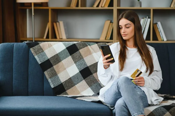 Happy young woman holding phone and credit card sitting on the couch. Online shopping