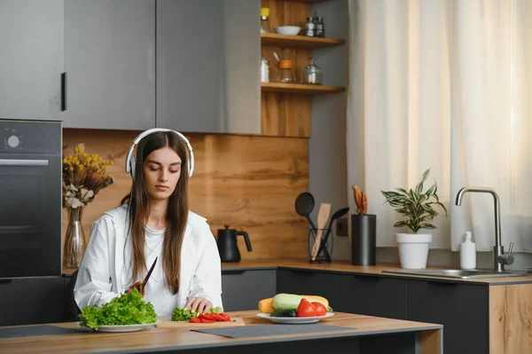 Adorable nice charming cheerful pretty beautiful stylish excited girl listening to music, singing in modern light white kitchen, cooking homemade dish with domestic products.