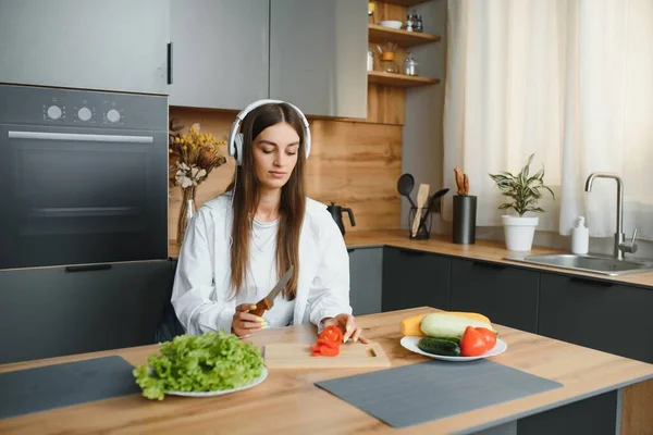 Adorable nice charming cheerful pretty beautiful stylish excited girl listening to music, singing in modern light white kitchen, cooking homemade dish with domestic products.