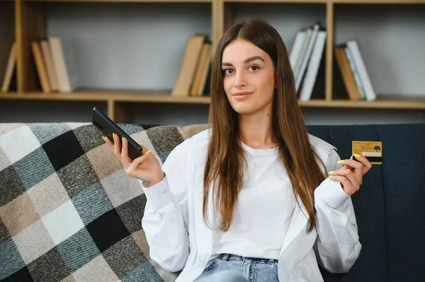 Happy young woman holding phone and credit card sitting on the couch. Online shopping