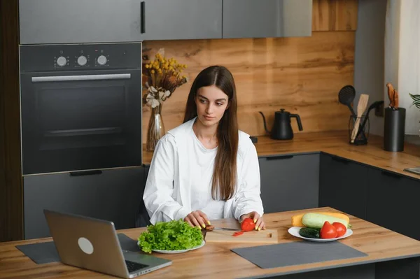 Athletic woman blogger nutritionist prepare a salad with fresh vegetables and conducts a video conference on healthy eating on laptop in the kitchen.