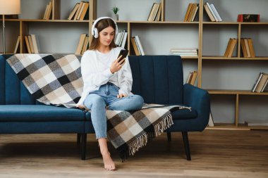 Portrait of attractive girl 20s wearing headphones using smartphone and listening to music while sitting on sofa at home.