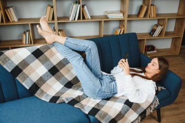 Young beautiful woman wearing white t-shirt on textile sofa at home. Attractive slim female in domestic situation, resting on couch in her lofty apartment. Background, copy space, close up