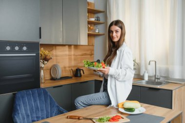 Mutlu gülümseyen tatlı kadın, evde bir sürü sebzeyle taze ve sağlıklı bir vegan salatası hazırlıyor ve yeni bir tarif deniyor..
