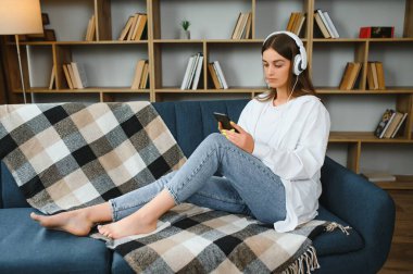 Beautiful young caucasian woman in casual clothing enjoying music and smiling while resting at home. Young woman with headphones using laptop at home.