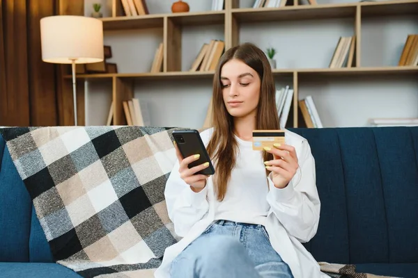 Happy young woman holding phone and credit card sitting on the couch. Online shopping
