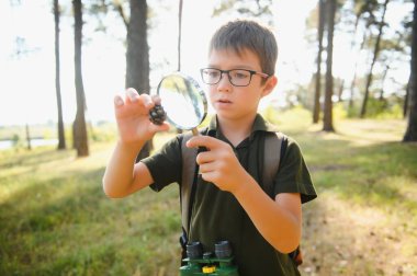 schoolboy is exploring nature with magnifying glass. Summer vacation for inquisitive kids in forest. Hiking. Boy-scout