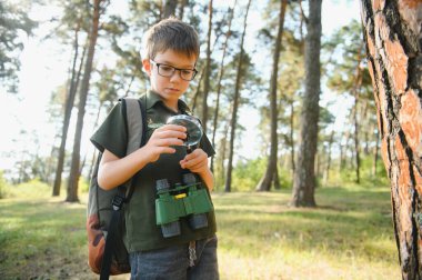 schoolboy is exploring nature with magnifying glass. Little child is looking on leaf of fern with magnifier. Summer vacation for inquisitive kids in forest. Hiking. Boy-scout