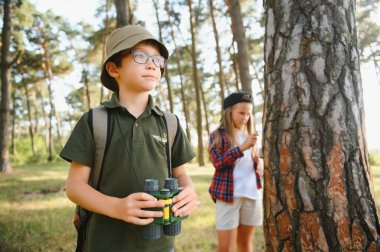 Two happy children having fun during forest hike on beautiful day in pine forest. Cute boy scout with binoculars during hiking in summer forest. Concepts of adventure, scouting and hiking tourism.