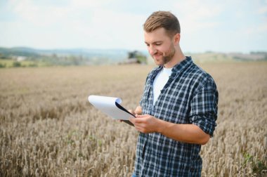 Happy farmer proudly standing in a field. Combine harvester driver going to crop rich wheat harvest. Agronomist wearing flannel shirt, looking at camera on a farmland.