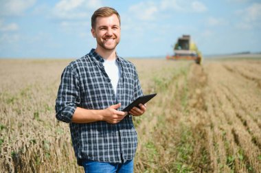 Happy farmer proudly standing in a field. Combine harvester driver going to crop rich wheat harvest. Agronomist wearing flannel shirt, looking at camera on a farmland.