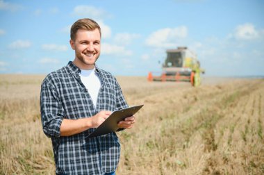 Happy farmer proudly standing in a field. Combine harvester driver going to crop rich wheat harvest. Agronomist wearing flannel shirt, looking at camera on a farmland.