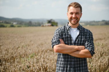Happy farmer proudly standing in a field. Combine harvester driver going to crop rich wheat harvest. Agronomist wearing flannel shirt, looking at camera on a farmland.