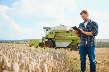 Happy farmer proudly standing in a field. Combine harvester driver going to crop rich wheat harvest. Agronomist wearing flannel shirt, looking at camera on a farmland.