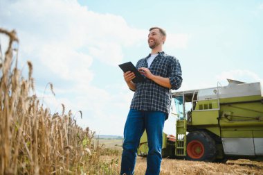 Happy farmer proudly standing in a field. Combine harvester driver going to crop rich wheat harvest. Agronomist wearing flannel shirt, looking at camera on a farmland.