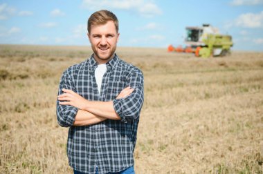 Happy farmer proudly standing in a field. Combine harvester driver going to crop rich wheat harvest. Agronomist wearing flannel shirt, looking at camera on a farmland.
