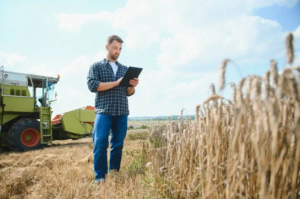 Handsome farmer with tablet standing in front of combine harvester during harvest in field