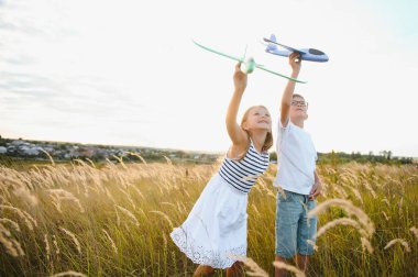 Running boy and girl holding two green and blue airplanes toy in the field during summer sunny day.