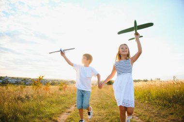 Running boy and girl holding two green and blue airplanes toy in the field during summer sunny day.