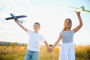 Running boy and girl holding two green and blue airplanes toy in the field during summer sunny day.