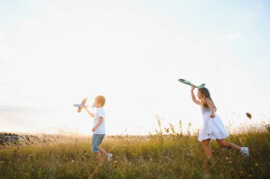 Running boy and girl holding two green and blue airplanes toy in the field during summer sunny day.