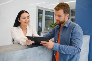 The employee of the beauty salon meets the client in the reception of a modern beauty salon. A man signs a paper with the consent for maintenance. The woman smiles at him.