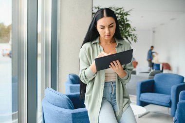 Young modern female freelancer in airport lounge or business center and works using digital tablet, creating new design project.