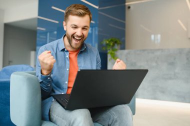 happy smiling remote online working man in casual outfit with laptop in joyful successful winning gesture sitting in an coworking office at a work desk