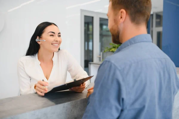 The employee of the beauty salon meets the client in the reception of a modern beauty salon. A man signs a paper with the consent for maintenance. The woman smiles at him.