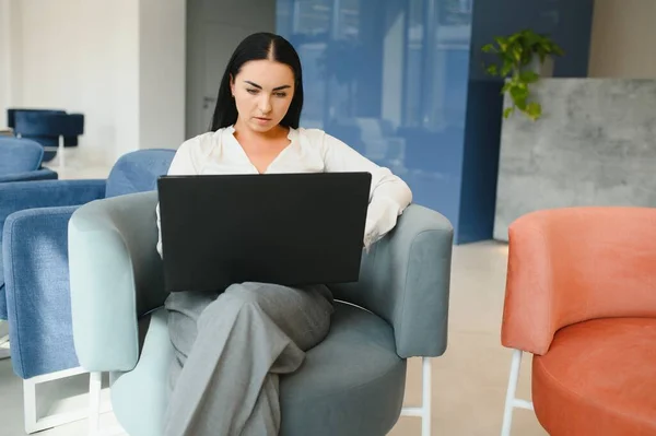 Young business woman sitting at sofa and working with laptop at the waiting hall of the business center.