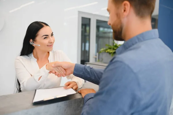 The employee of the beauty salon meets the client in the reception of a modern beauty salon. A man signs a paper with the consent for maintenance. The woman smiles at him.