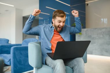happy smiling remote online working man in casual outfit with laptop in joyful successful winning gesture sitting in an coworking office at a work desk