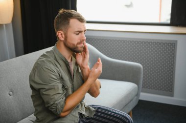 Diş sağlığı sorunları konsepti. Güçlü diş ağrısı çeken bir adam. İltihaplı sinirleri, diş apsesi ya da diş hassasiyeti olan üzgün bir erkek hasta kanepeye oturmuş ve acı içinde yanaklarına dokunuyor.
