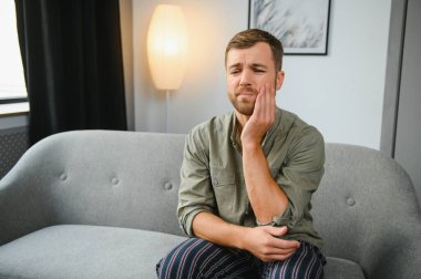 Problems with teeth. Toothache. Upset caucasian guy is sitting on the couch at home, holding his hand to his cheek, having a toothache, needs a dentist consultation, sad facial expression