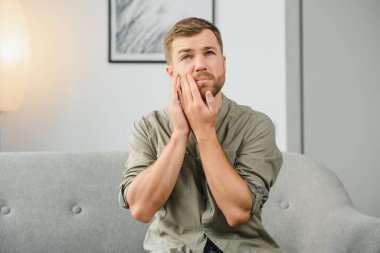 Problems with teeth. Toothache. Upset caucasian guy is sitting on the couch at home, holding his hand to his cheek, having a toothache, needs a dentist consultation, sad facial expression