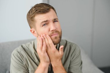 Diş sağlığı sorunları konsepti. Güçlü diş ağrısı çeken bir adam. İltihaplı sinirleri, diş apsesi ya da diş hassasiyeti olan üzgün bir erkek hasta kanepeye oturmuş ve acı içinde yanaklarına dokunuyor.