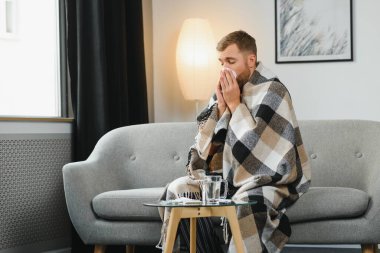Kötü soğuk algınlığı ya da mevsimsel grip virüsü taşıyan hasta yetişkin bir adam evde hastalıkla savaşıyor. Mutsuz orta yaşlı bir adam sıcak atkı ve ekoseye sarılı olarak koltukta oturuyor, kağıt mendil tutuyor ve hapşırıyor.