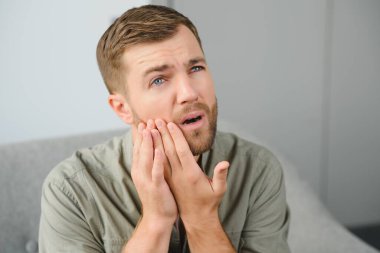 Diş sağlığı sorunları konsepti. Güçlü diş ağrısı çeken bir adam. İltihaplı sinirleri, diş apsesi ya da diş hassasiyeti olan üzgün bir erkek hasta kanepeye oturmuş ve acı içinde yanaklarına dokunuyor.