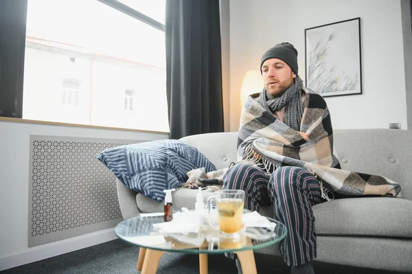 It's cold at home in wintertime. Man freezing in his house in winter because of broken thermostat. young guy wrapped in woolen plaid shivering while sitting on sofa in living room interior.