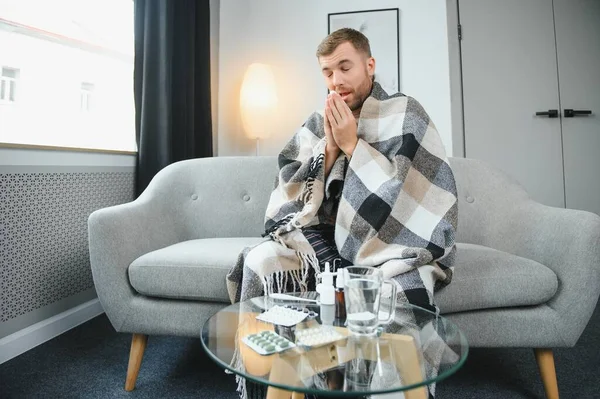 Young man suffering from a common cold and flu or allergy sit at home wrapped in blanket and wipe his nose with tissues while he have a strong headache pain, healthcare concept