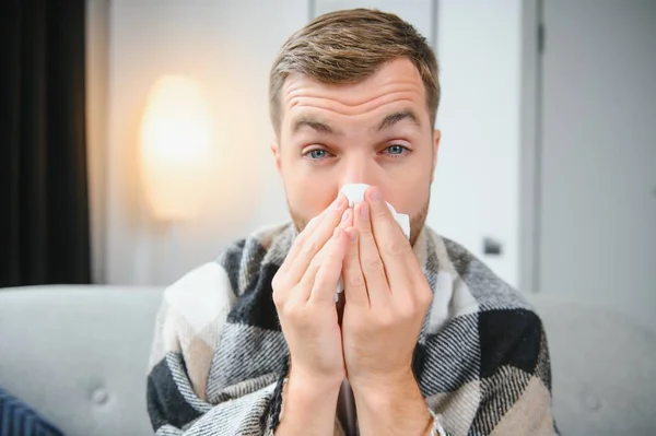 Young man suffering from a common cold and flu or allergy sit at home wrapped in blanket and wipe his nose with tissues while he have a strong headache pain, healthcare concept