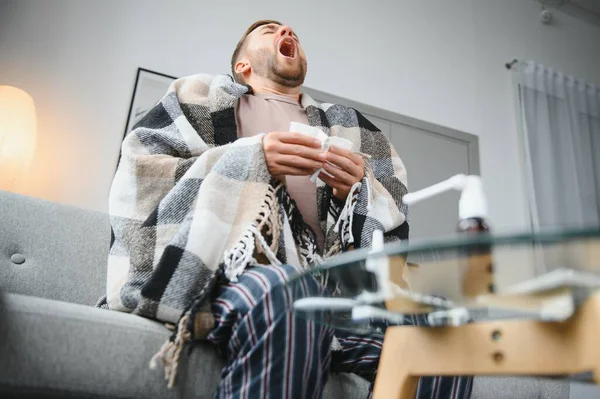 Young man suffering from a common cold and flu or allergy sit at home wrapped in blanket and wipe his nose with tissues while he have a strong headache pain, healthcare concept