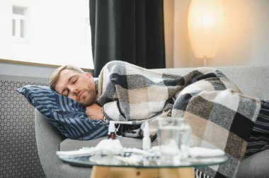 Hasta sakallı adam, soğuk algınlığı ya da mevsimsel grip olmuş, evdeki kanepede oturuyor. Ateşli, ekoseli ve endişeli yüzüyle titreyen bir adam.