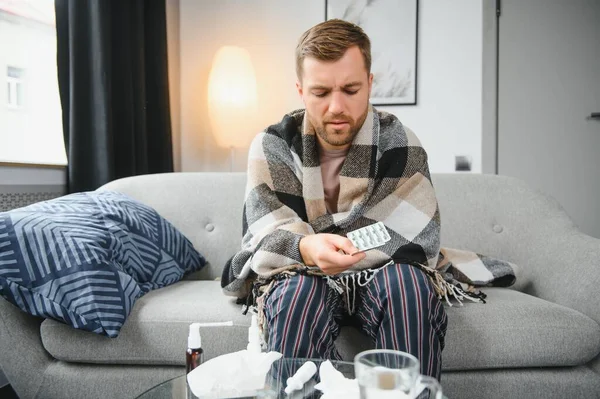 It's cold at home in wintertime. Man freezing in his house in winter because of broken thermostat. young guy wrapped in woolen plaid shivering while sitting on sofa in living room interior.