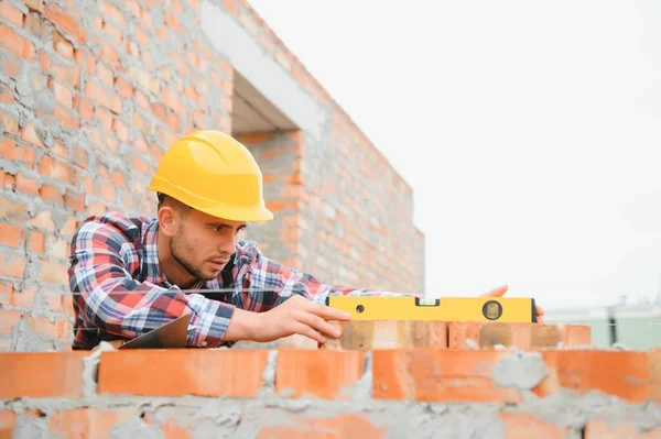 Using bricks. Young construction worker in uniform is busy at the unfinished building.