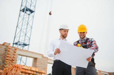 İnşaat mühendisleri inşaat projesinin ilerlemesini denetliyor. Yeni beton zemin üstü çatı ve vinç arka planı üzerinde duruyorlar.