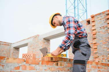 Tuğla duvarla meşgul. Üniformalı inşaat işçisi ve güvenlik ekipmanı inşaatında çalışıyor