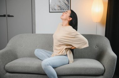 Sırt ağrısı çeken olgun bir kadın oturma odasındaki koltukta oturan mutsuz, yaşlı bir sarışın sırt ağrısı yüzünden rahatsız..