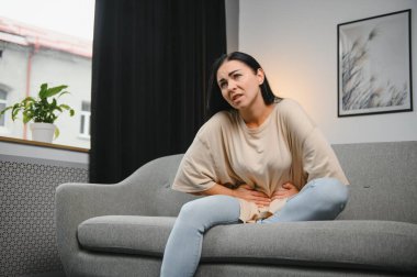 Genç bir kadın evdeki kanepede otururken karın ağrısı çekiyor. Karın ağrısının nedeni adet sancısı, mide iltihabı, mide ülseri, gıda zehirlenmesi, ishal ya da bağırsak iltihabı. Seçici odak.